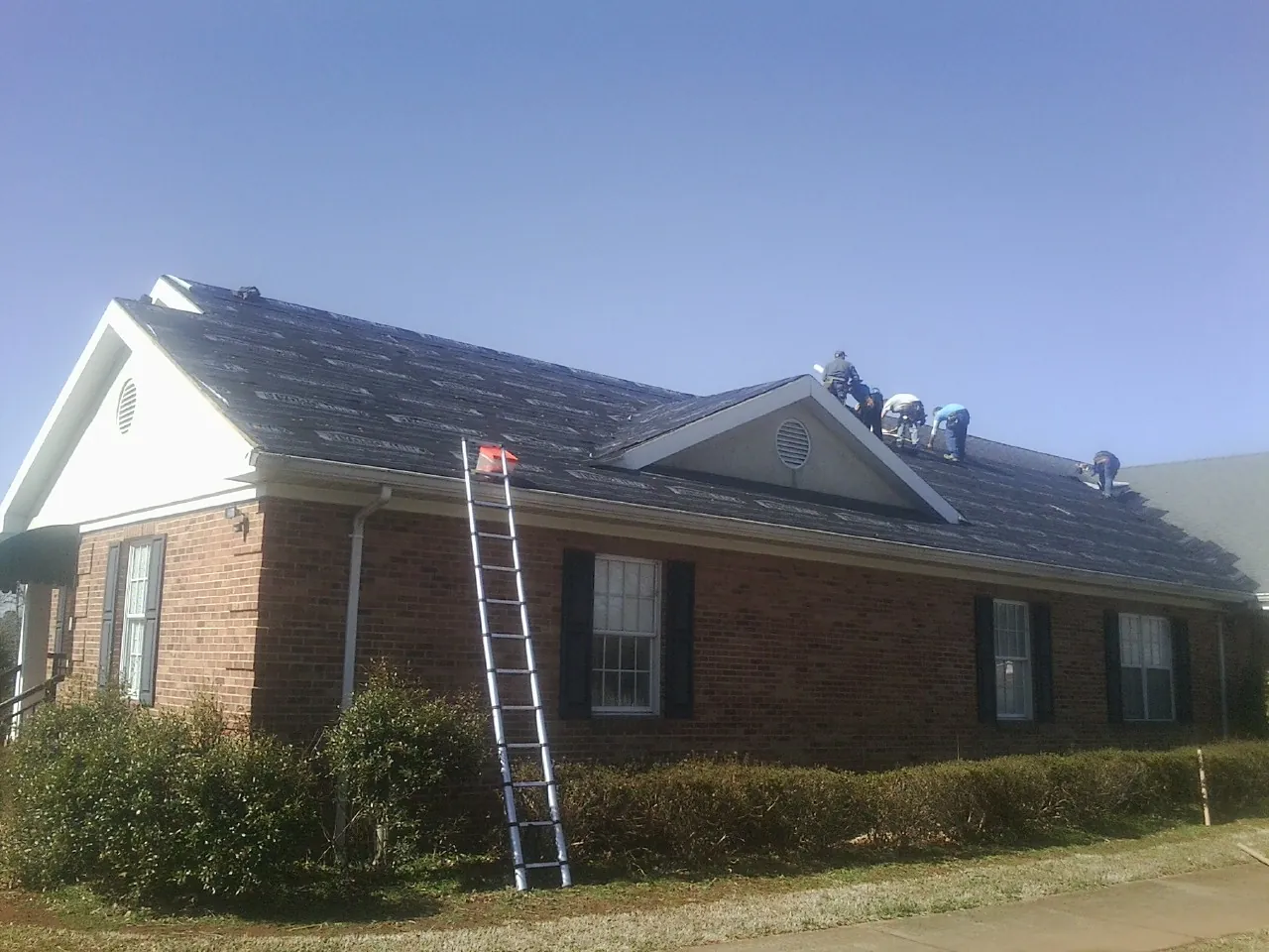 Crew tearing shingles off a brick ranch roof with a ladder set — Metal Roof Repair work by our crew in Claypool