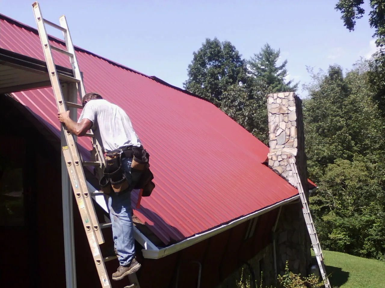 Roofer on a ladder beside a red metal roof and stone chimney — Soffit & Fascia Repair by Roofing Brothers in Claypool